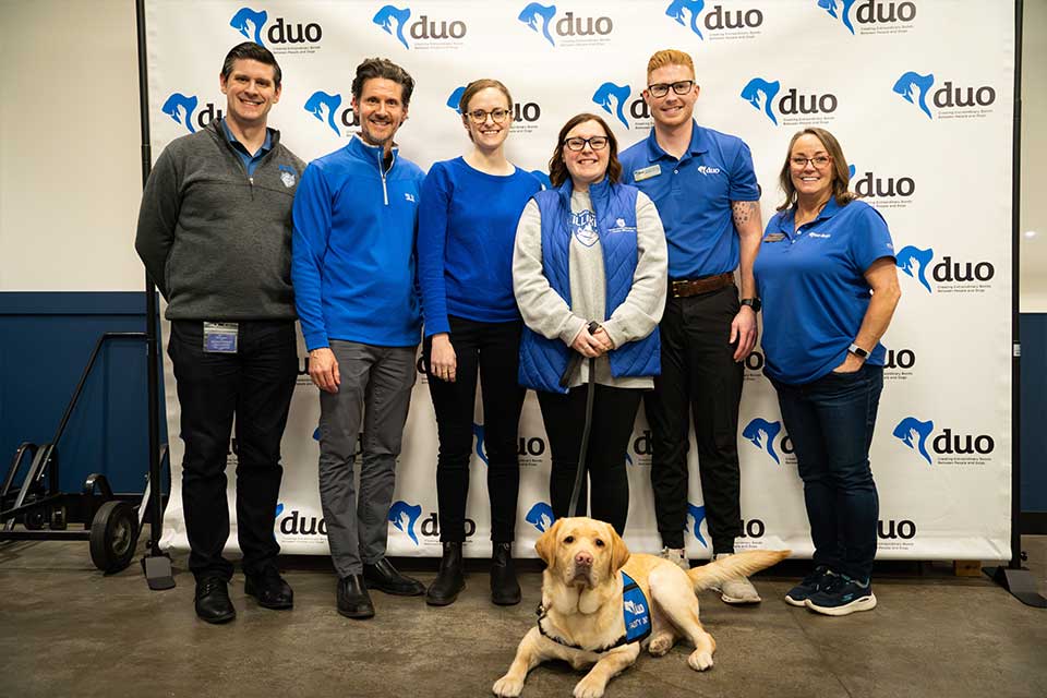 Woody, an yellow retriever, lays on the ground as his six handlers stand behind him at his graduation to become SLU's facility dog. 